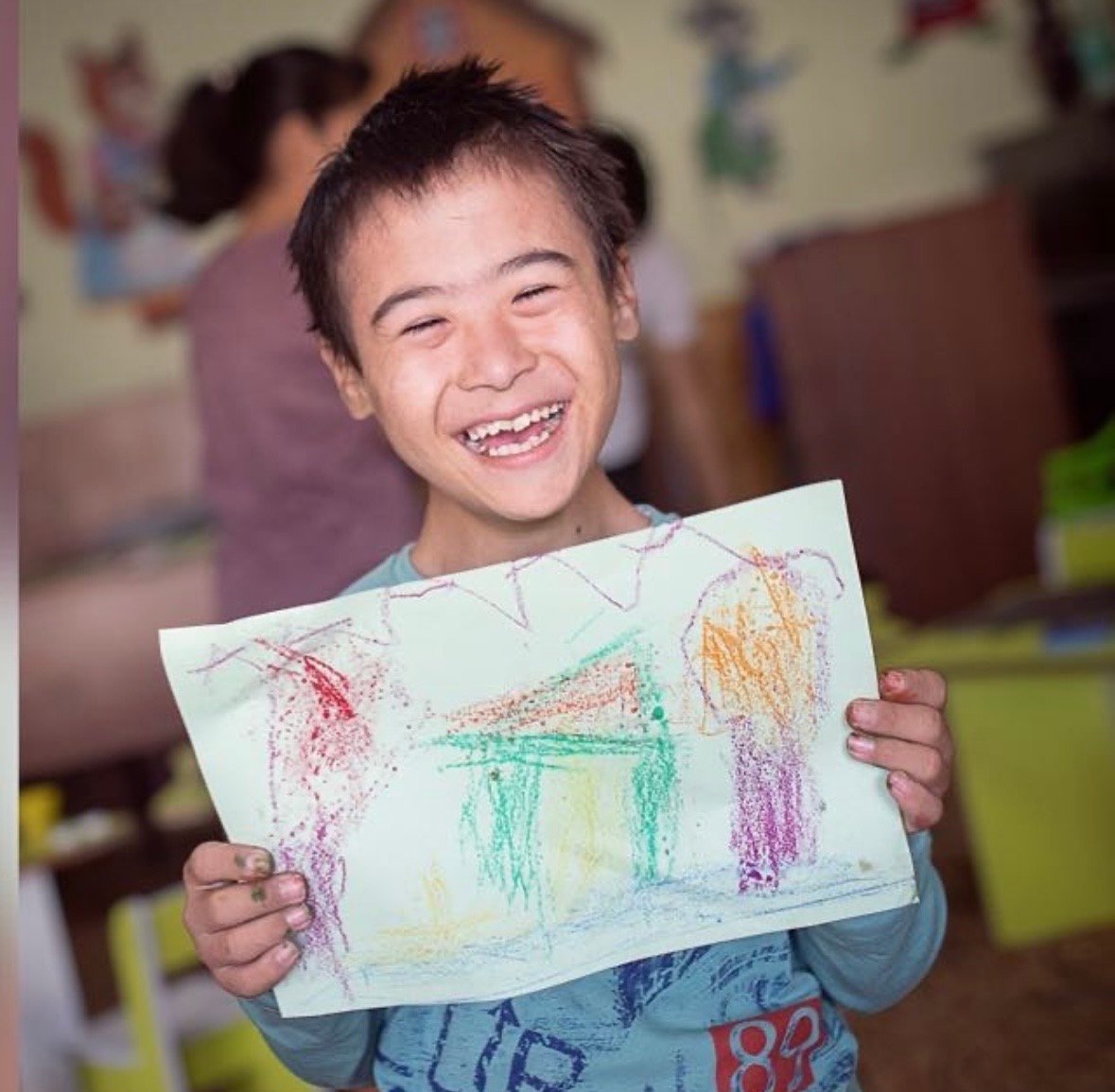 A laughing boy with Down syndrome holds up his colourful crayon drawing