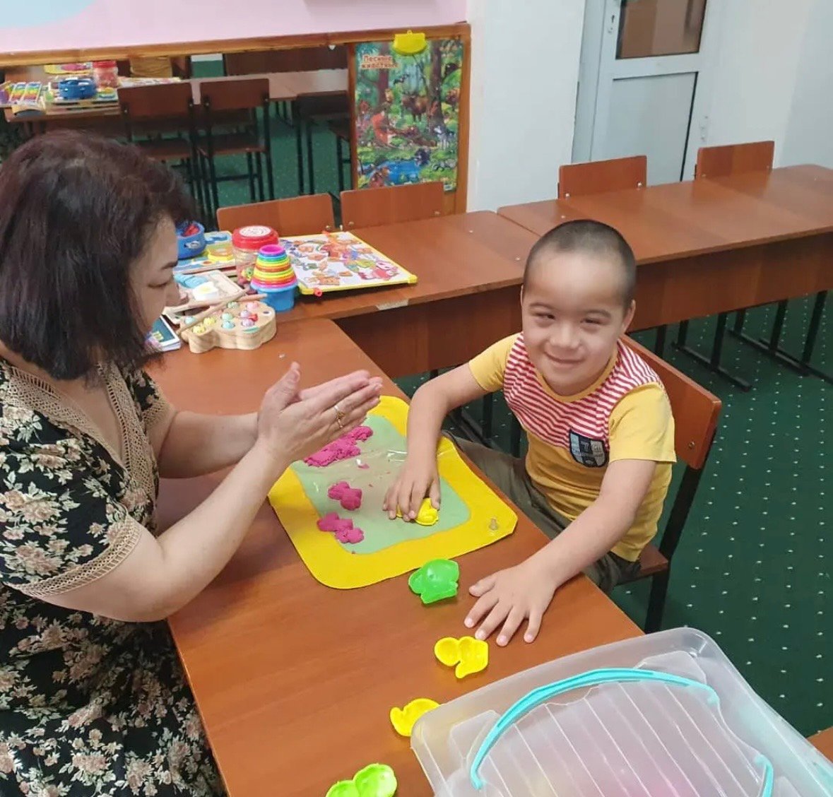 A teacher claps as a smiling boy with Down syndrome shapes modelling clay at a classroom table